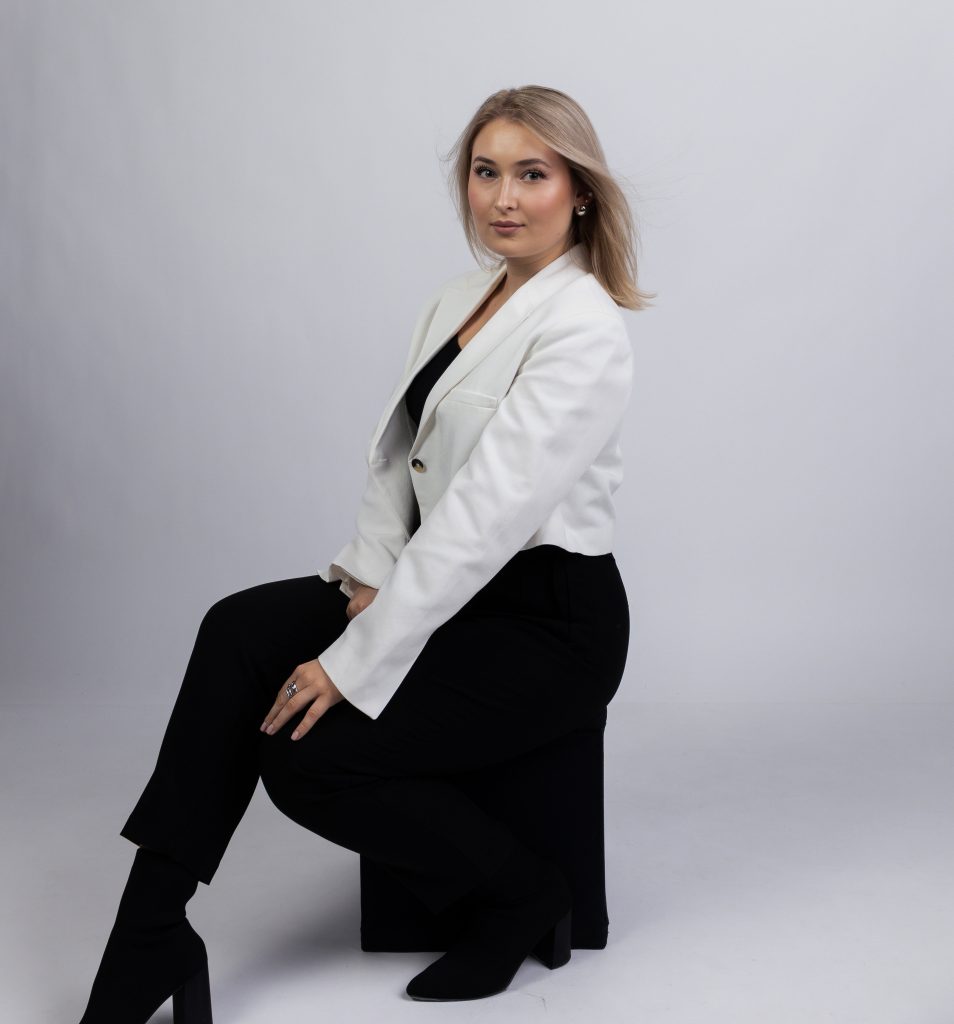 Kobieta o blond włosach, ubrana w białą marynarkę i czarne spodnie, siedzi na czarnej pufie na jasnym, jednolitym tle. Jest zwrócona bokiem do obiektywu, ale patrzy prosto w kamerę z lekkim uśmiechem.