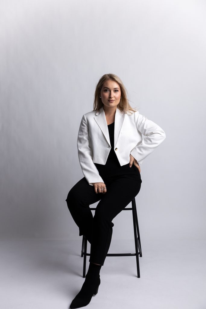 Kobieta w blond włosach siedzi przodem na czarnym hokerze na jasnoszarym tle. Ma na sobie krótką białą marynarkę, czarną bluzkę i czarne spodnie. Lewą rękę opiera na biodrze, patrząc pewnie w stronę aparatu.